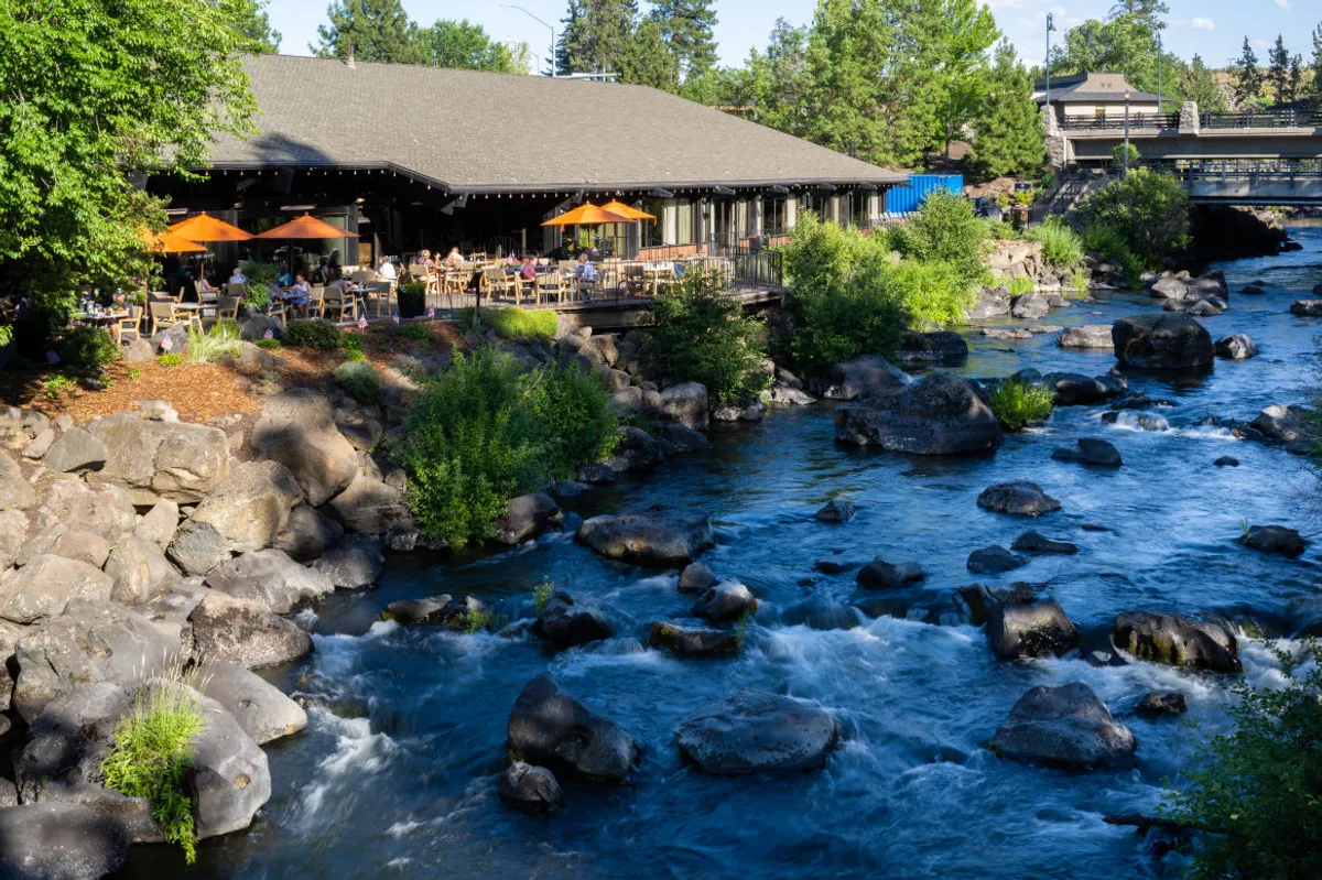 The image showcases an inviting restaurant patio with vibrant orange umbrellas overlooking the tranquil Deschutes River, surrounded by natural rocks and lush trees. The setting is bathed in the warm glow of sunset, suggesting a relaxed and cheerful atmosphere where guests can dine al fresco, enjoy live music, and soak in the scenic beauty of Central Oregon.