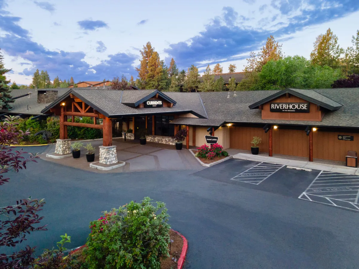 This exterior image showcases the main entrance to the property, prominently featuring signage for both Currents Restaurant and Riverhouse. The view also includes meticulously landscaped gardens, hinting at a serene atmosphere, and captures guests dining at Currents, relaxing in the garden, and socializing in the nearby lobby area.