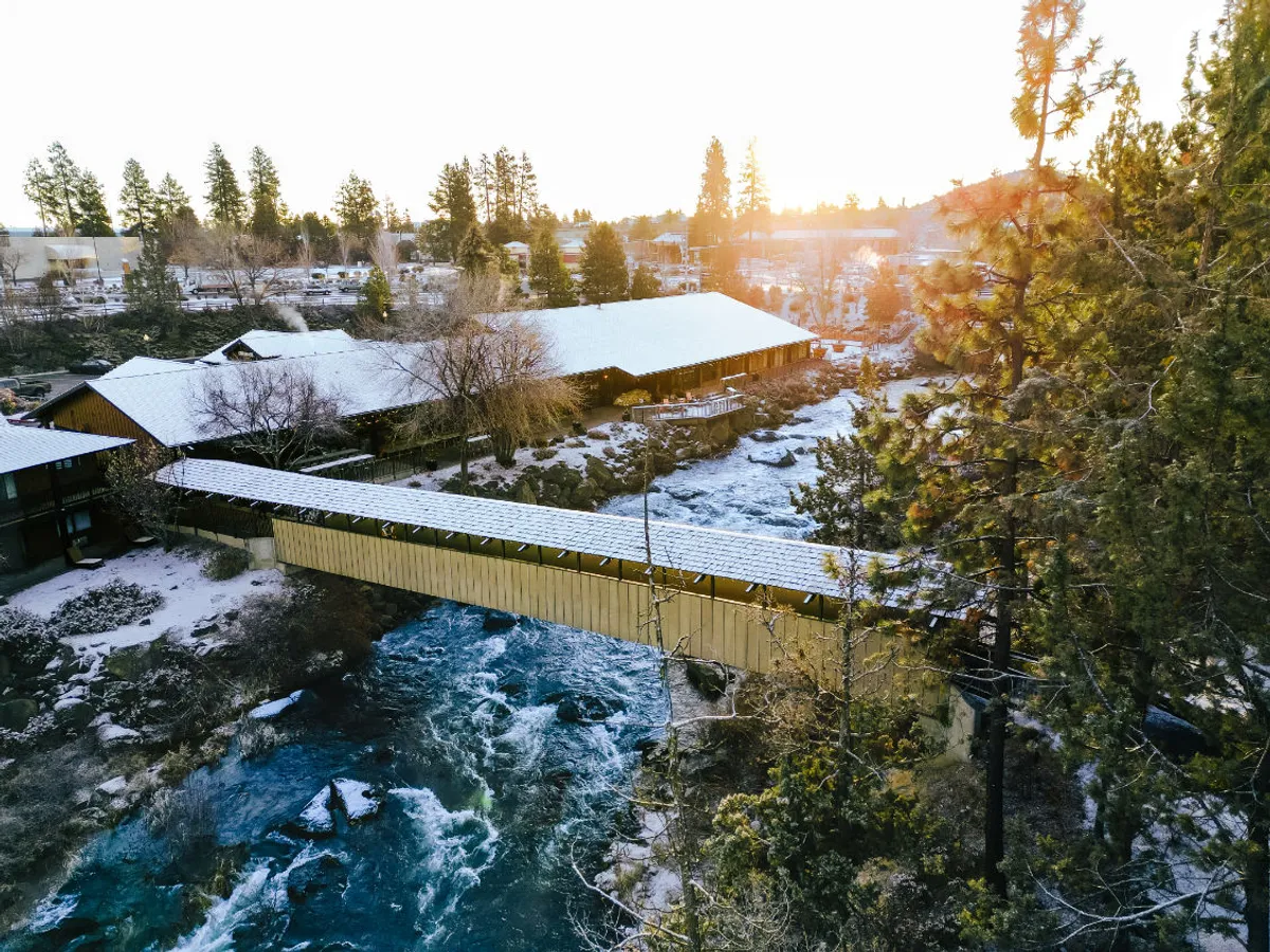 This image captures a picturesque covered bridge spanning the flowing Deschutes River, framed by lush trees and with snow-dusted roofs visible. The scene is tranquil and welcoming, inviting exploration and outdoor enjoyment. The bridge connects walking trails and hints at the natural adventures that await just beyond the lodge.