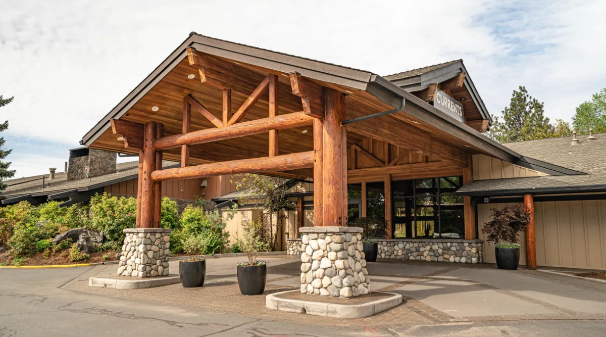An exterior perspective of the property highlighting architectural features such as large wooden beams, sturdy stone pillars, expansive windows, and lush surrounding greenery. Arriving guests and greeting staff are visible, emphasizing a welcoming vibe and connection between indoors and outdoors.