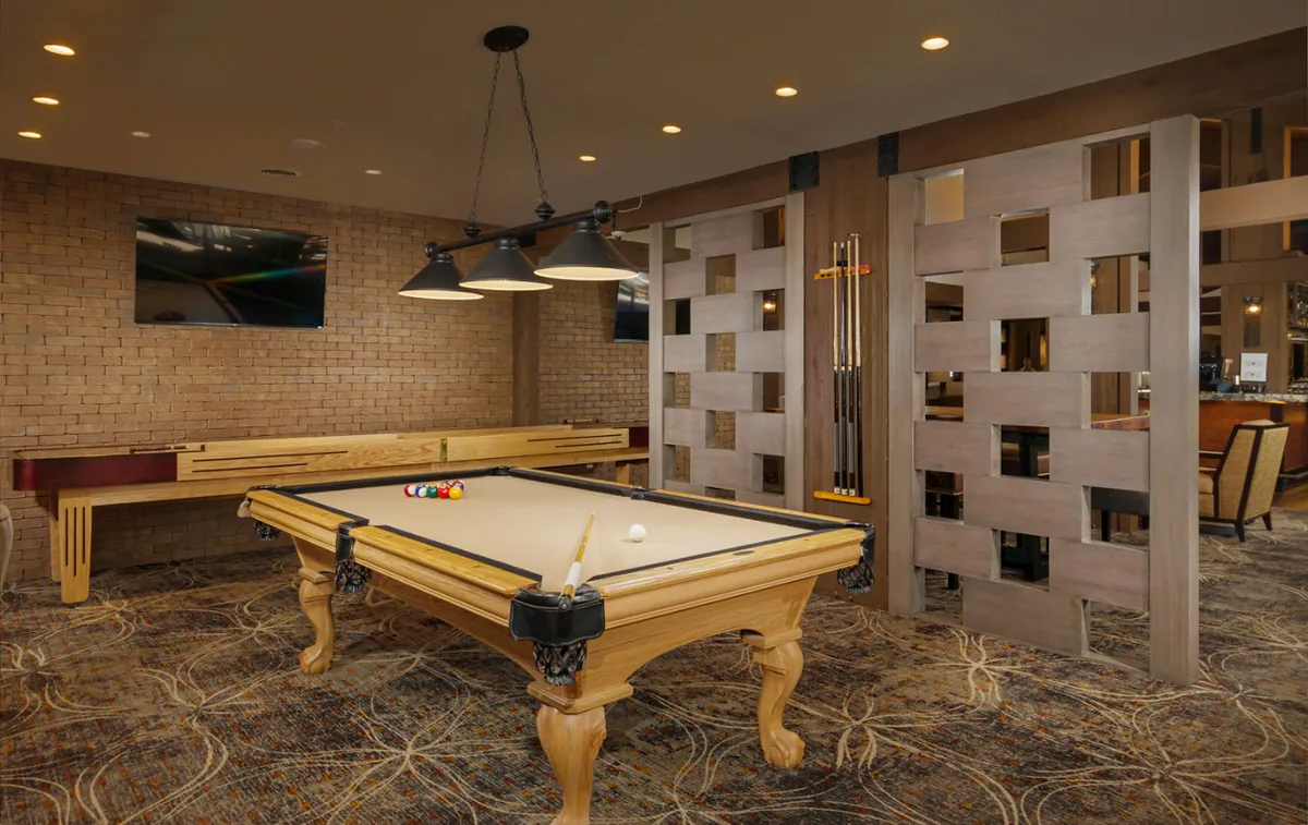 This interior shot centers on a pool table with stylish pendant lighting above and textured wall panels behind, evoking a relaxed, fun mood. The space clearly encourages friendly competitions and group activities, with the TV in view hinting at sports entertainment.
