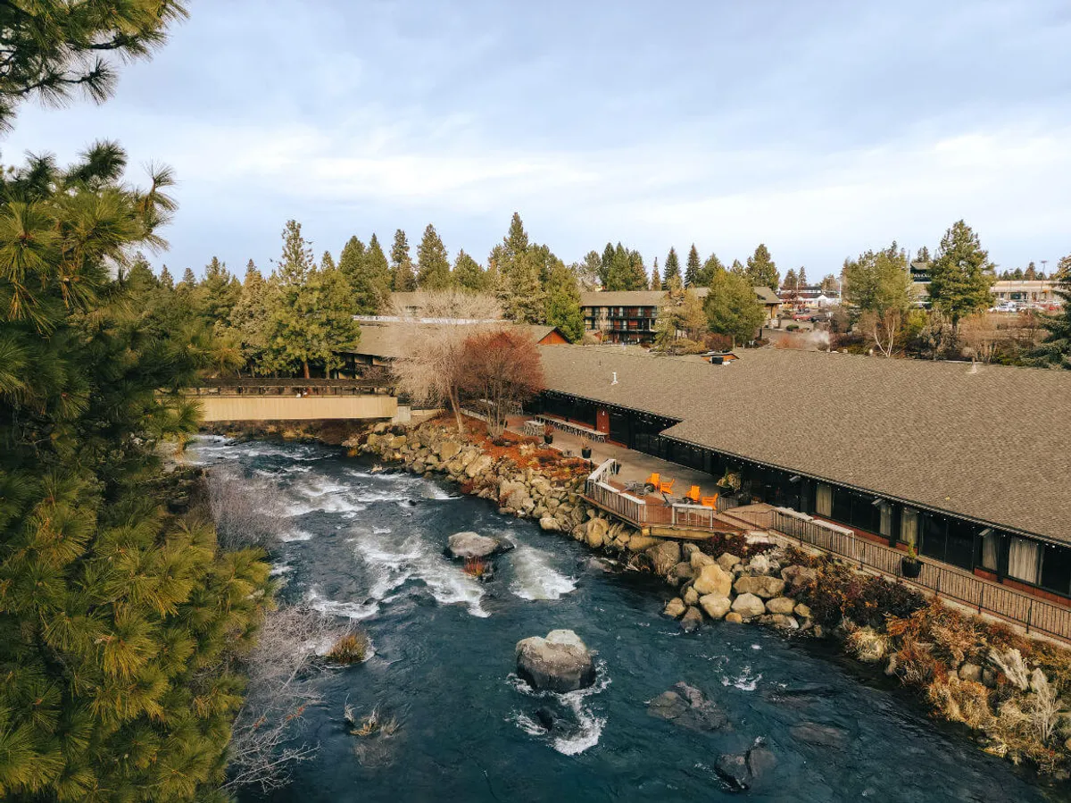 This image features a scenic riverside with a building nestled among surrounding trees and a deck area overlooking the water. The setting evokes a tranquil atmosphere and hints at the possibility of enjoying nature from a private outdoor space, such as a balcony or patio. Sunset light warms the scene, emphasizing peaceful relaxation and connection to the outdoors.