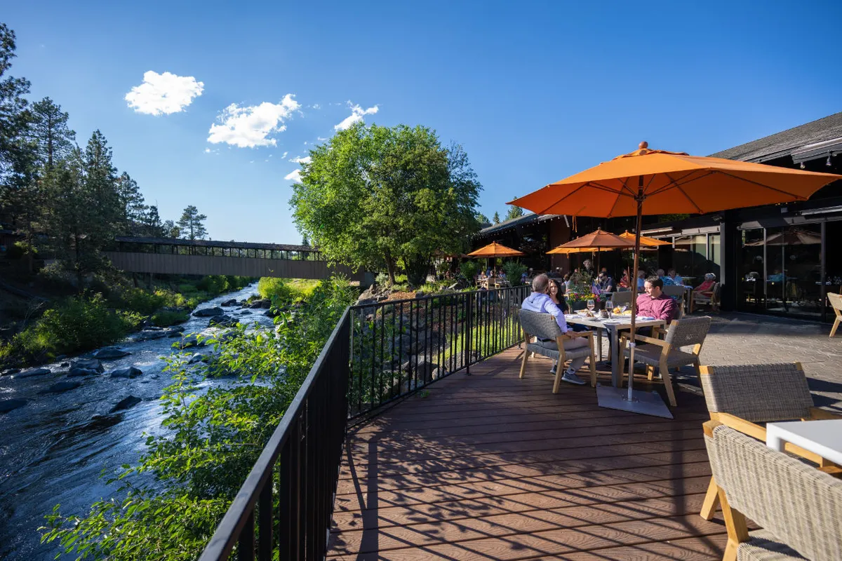 The image captures an outdoor seating area by the river, with orange umbrellas providing shade for guests enjoying dining and social moments. The scene is lively yet relaxed, with reflections of the sunset on the water enhancing the inviting mood. The river and lush surroundings evoke a sense of calm and natural beauty.