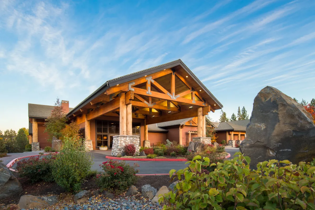 An exterior view of Riverhouse Lodge’s grand entrance, showcasing rustic wooden beams, stone walls, and beautifully landscaped gardens. The scene is bathed in golden sunset light, emphasizing a seamless blend between the lodge and its natural surroundings. Guests gather and relax outdoors, hinting at the start or end of an adventure-filled day.