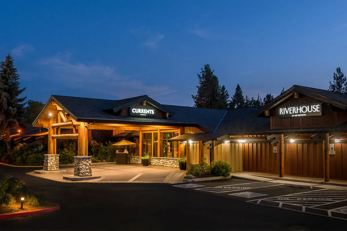 An inviting exterior view of CURRENTS’ main entrance, featuring wood accents, lush landscaping, and clear signage for both Currents and Riverhouse. Diners can be seen enjoying the alfresco experience under the stars, providing a sense of place and welcoming atmosphere.