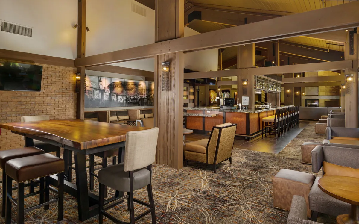 A spacious lounge area with a large wooden table, bar area, and comfortable seating arrangements, designed for relaxed socializing. The space is well-lit and features a welcoming, modern ambiance ideal for gatherings or informal meetings.