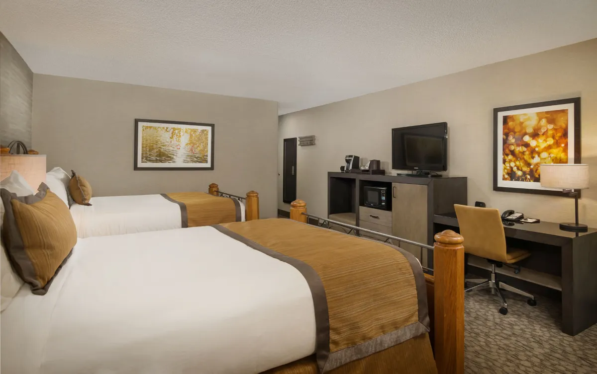 A cozy deluxe room featuring two queen beds with crisp linens, a functional work desk paired with a comfortable chair, a large TV unit set across from the beds, and tasteful framed artwork adorning the wall. The room is thoughtfully designed to blend relaxation with utility, and the spacious layout is ideal for couples or families.