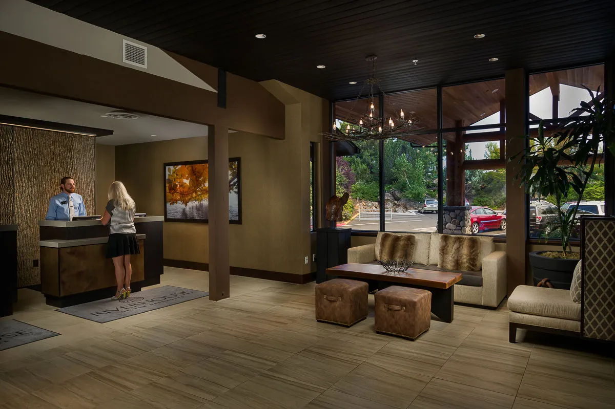 The image showcases the inviting reception area of Riverhouse Lodge, with guests checking in and relaxing in a comfortable seating area. Large windows frame views of the outdoors, bringing natural light and hints of the surrounding river landscape into the space. The scene feels warm and welcoming, emphasizing the beginning of a family getaway and the communal nature of the lodge.